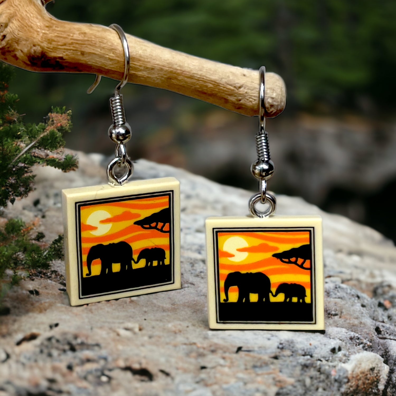 A pair of earrings with a lego tile printed with a elephant and her baby in silhouette with a sunrise on the background, hanging off a branch on a rock with a great blurred background 