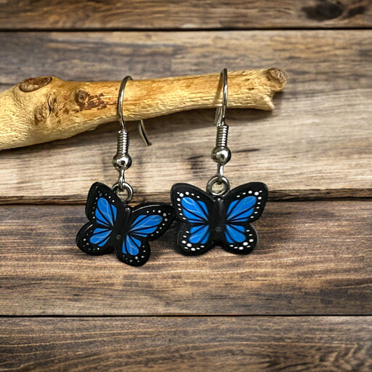 A pair of blue winged butterflies with a black outline made into a pair of silver hook earrings handing off a branch on a wooden background