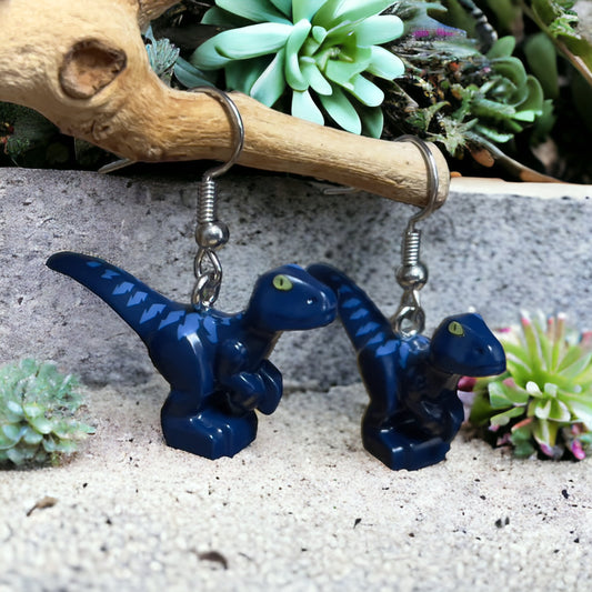 Two dark blue Lego t-rex style dinosaurs with light blue stripes down their backs as hook earrings. They are hanging off a branch above a grey rough textured surface with green succulent plants in the background.