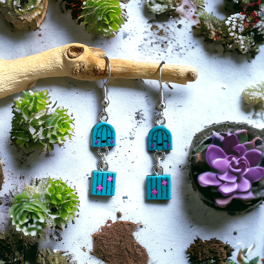 A cute set of little green cactus shaped lego earrings with pink stars and black outline made into a loop sytle earring with silver rings holding the top and bottom halves together hanging off a branch on a bed of succulents