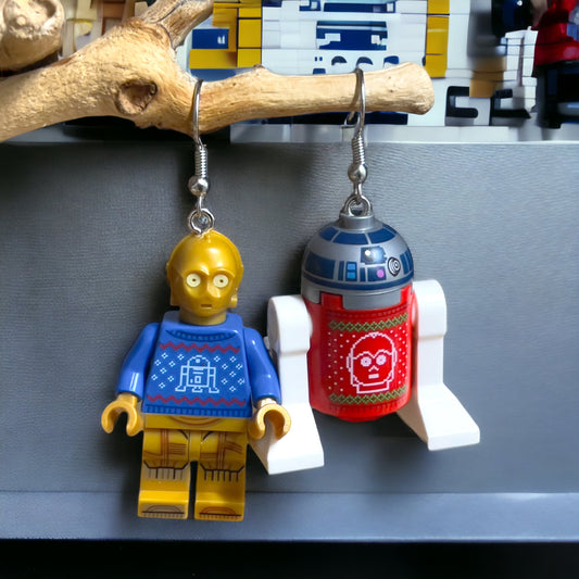 A lego C3PO who is gold with a blue christmas sweater with r2d2 on it and R2D2 with a red sweater on with c3po on it made into silver hook earrings hanging off a branch with a grey background