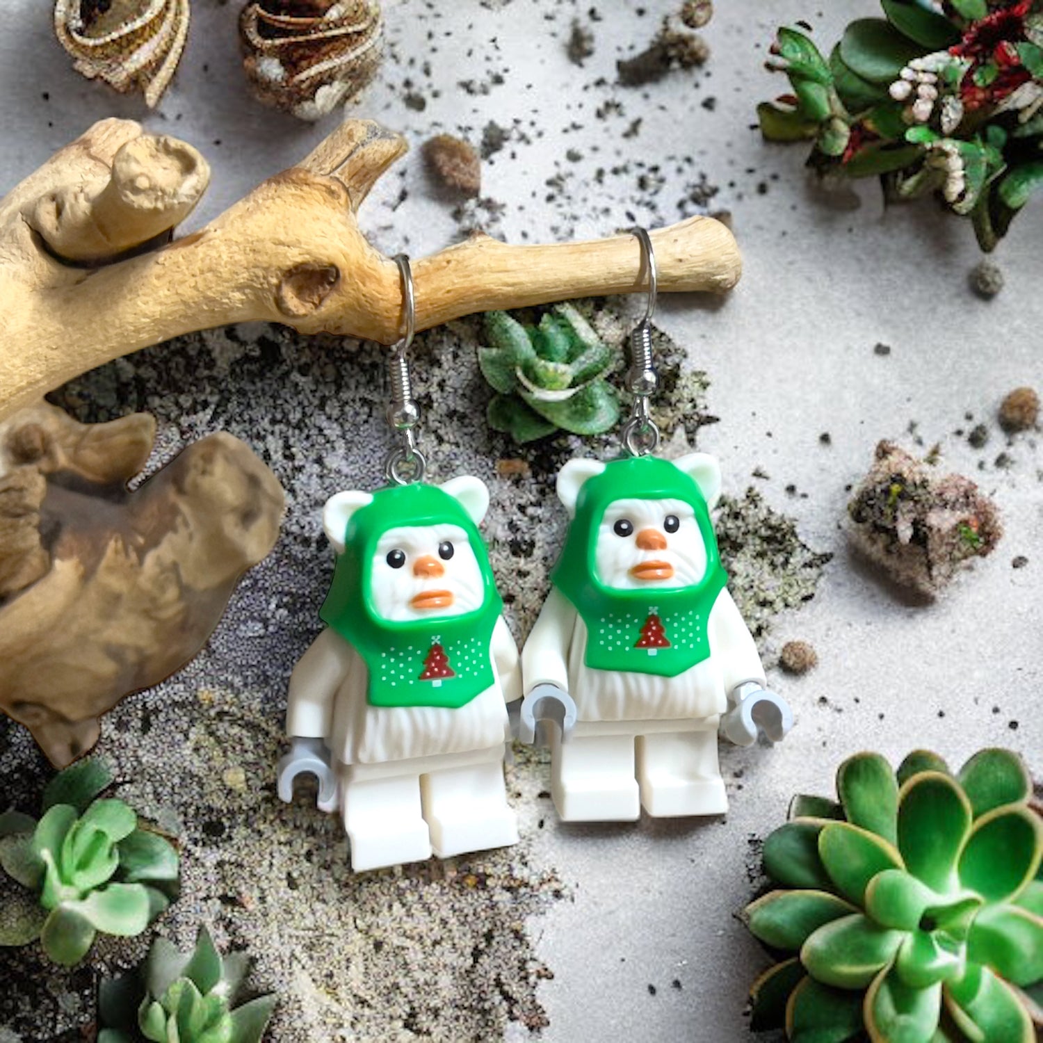 Two lego white ewoks with a green christmas hookie with silver hook earrings hanging on a branch with a background of succulents and sand.