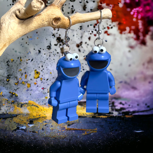 Two blue cookie monster lego minifigures with silver hook earrings hanging off a branch with a yellow, black and red paint splashed background