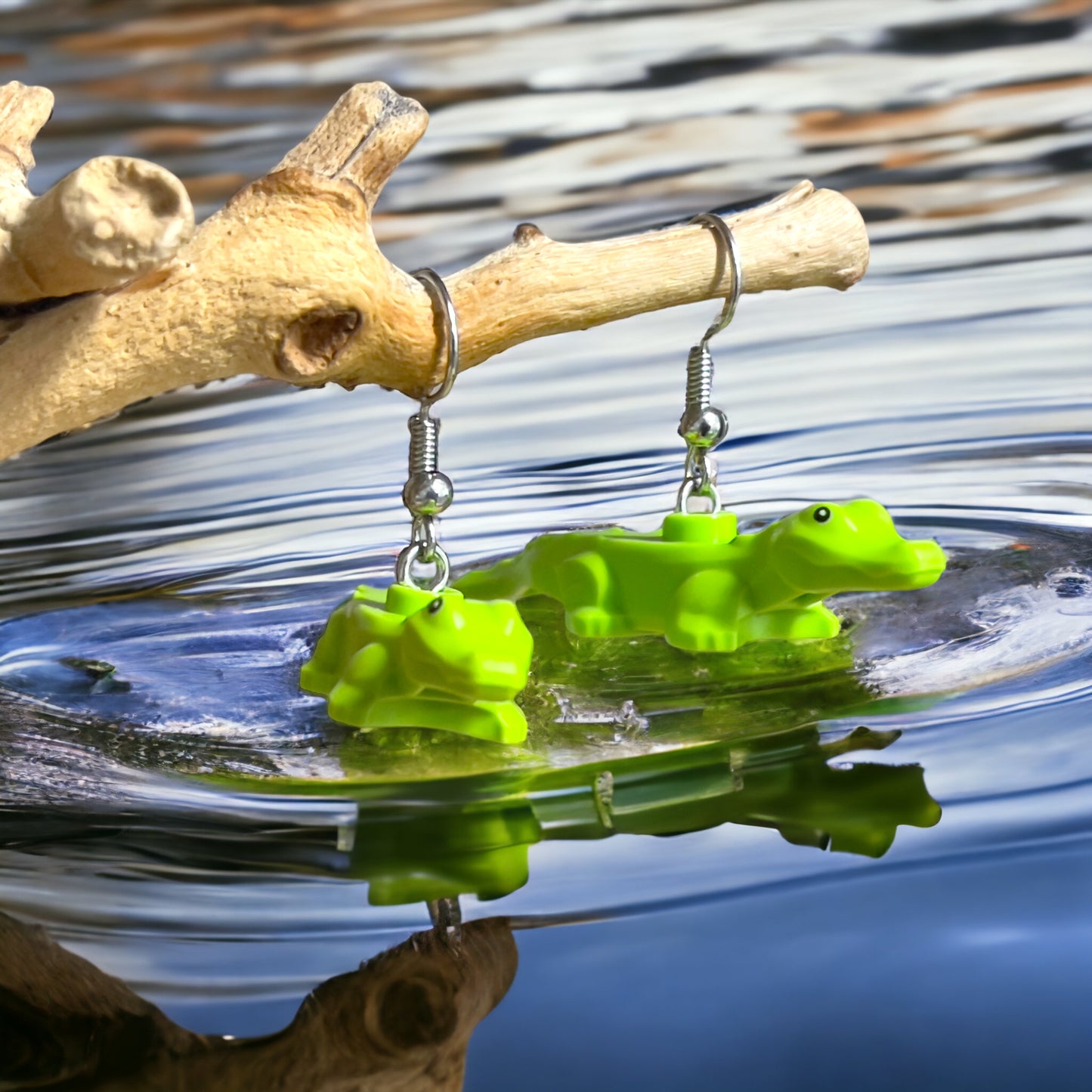 Two green baby lego crocodiles made into silver hook earrings hanging on a branch above water where they have made a ripple effect with their reflections in the water