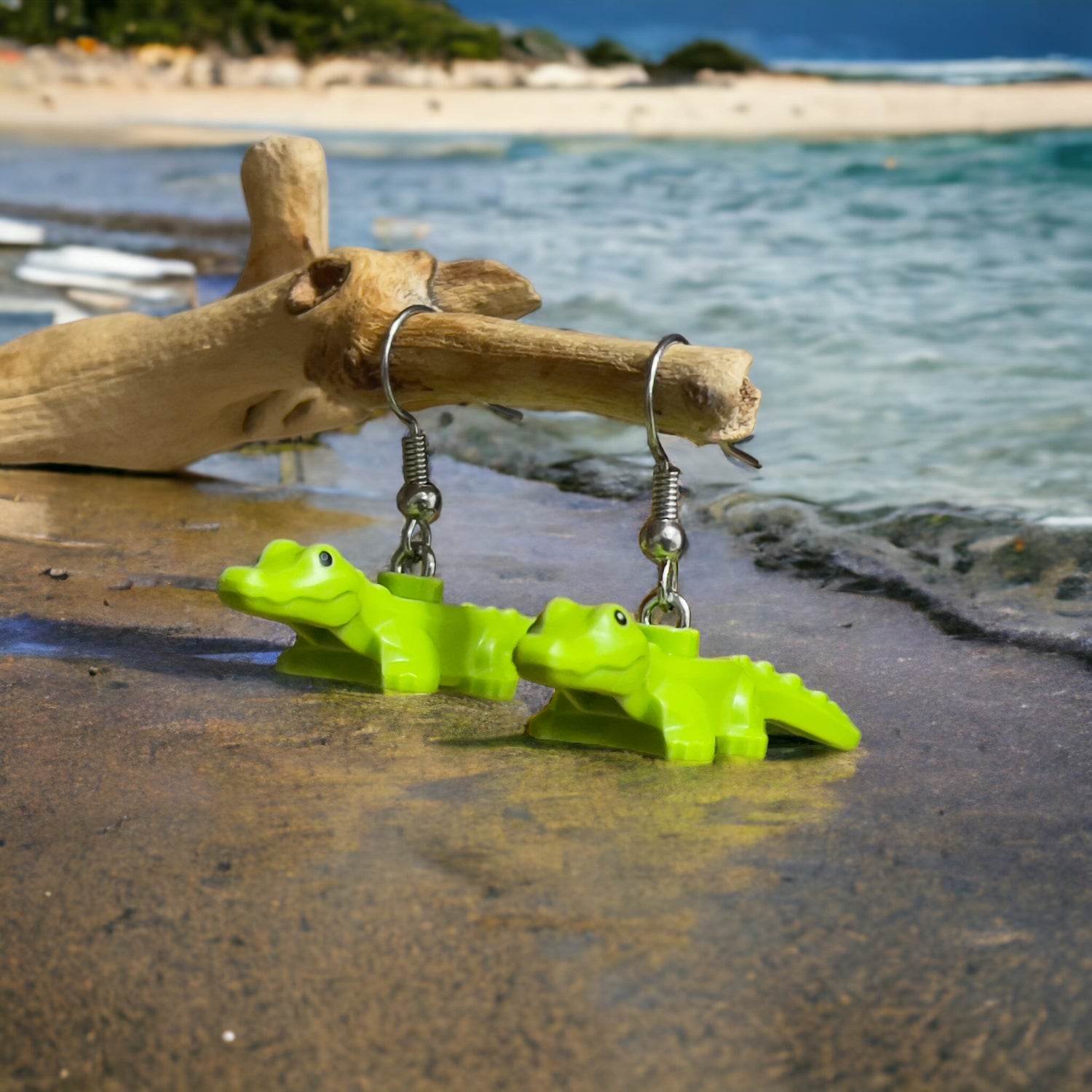 Two green lego baby crocodiles made into silver hook earrings hanging on a branch sitting on sand with water gently going over the sand with ocean waves in the background and a beach island in the far background