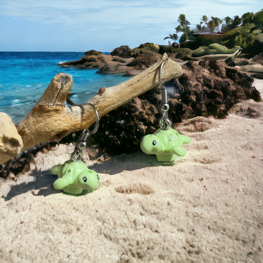 Two little light green lego turtles with shiny green spots and black eyes made into silver loop earrings hanging off a branch on sand with the blue ocean in the background and ricks and forest off to the top right of the pictures