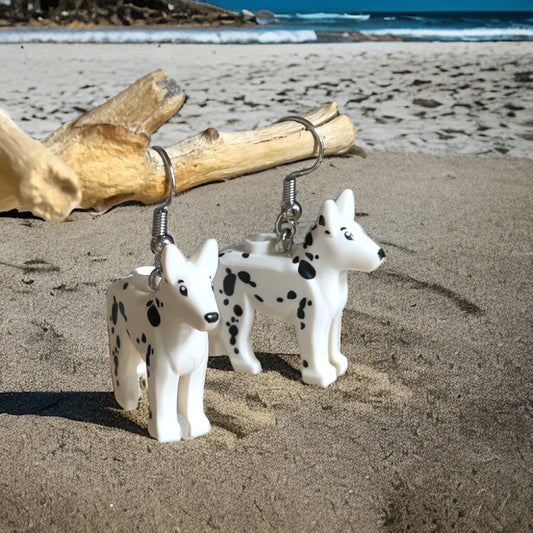 A set of two white dogs with black spots with hook silver earrings on a sandy beach scene with the water and waves in the background.