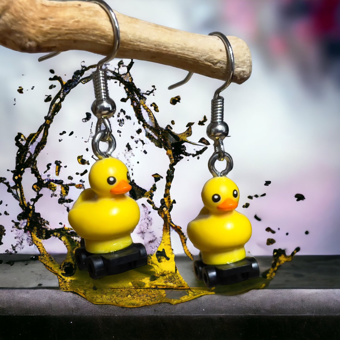 Two little yellow lego ducks on black lego roller skates with silver hook earrings hanging off a branch with a yellow paint splash underneath them with a white background and silver bench.