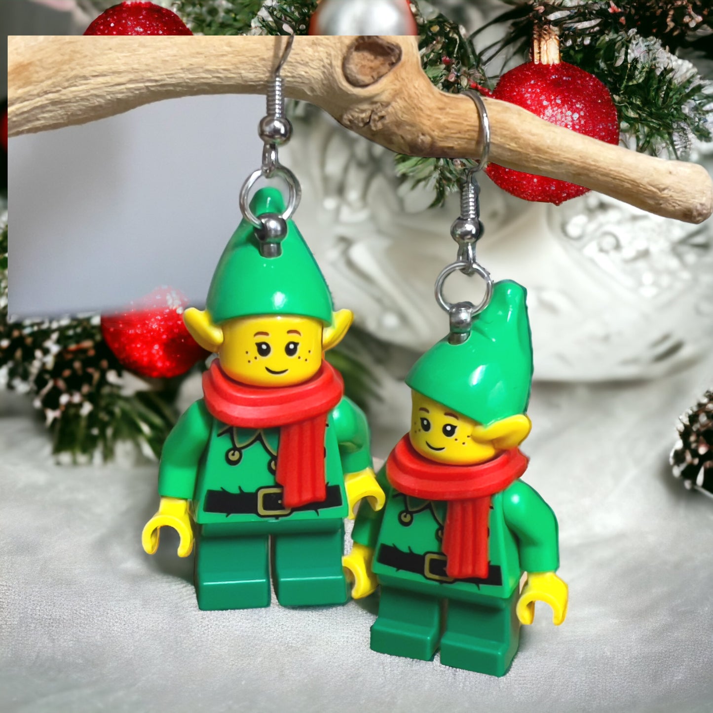 A lego minifigure of an Elf in a green suit a red scarf and green hat as hook earrings hanging off a branch in some green and white background and red round decorations