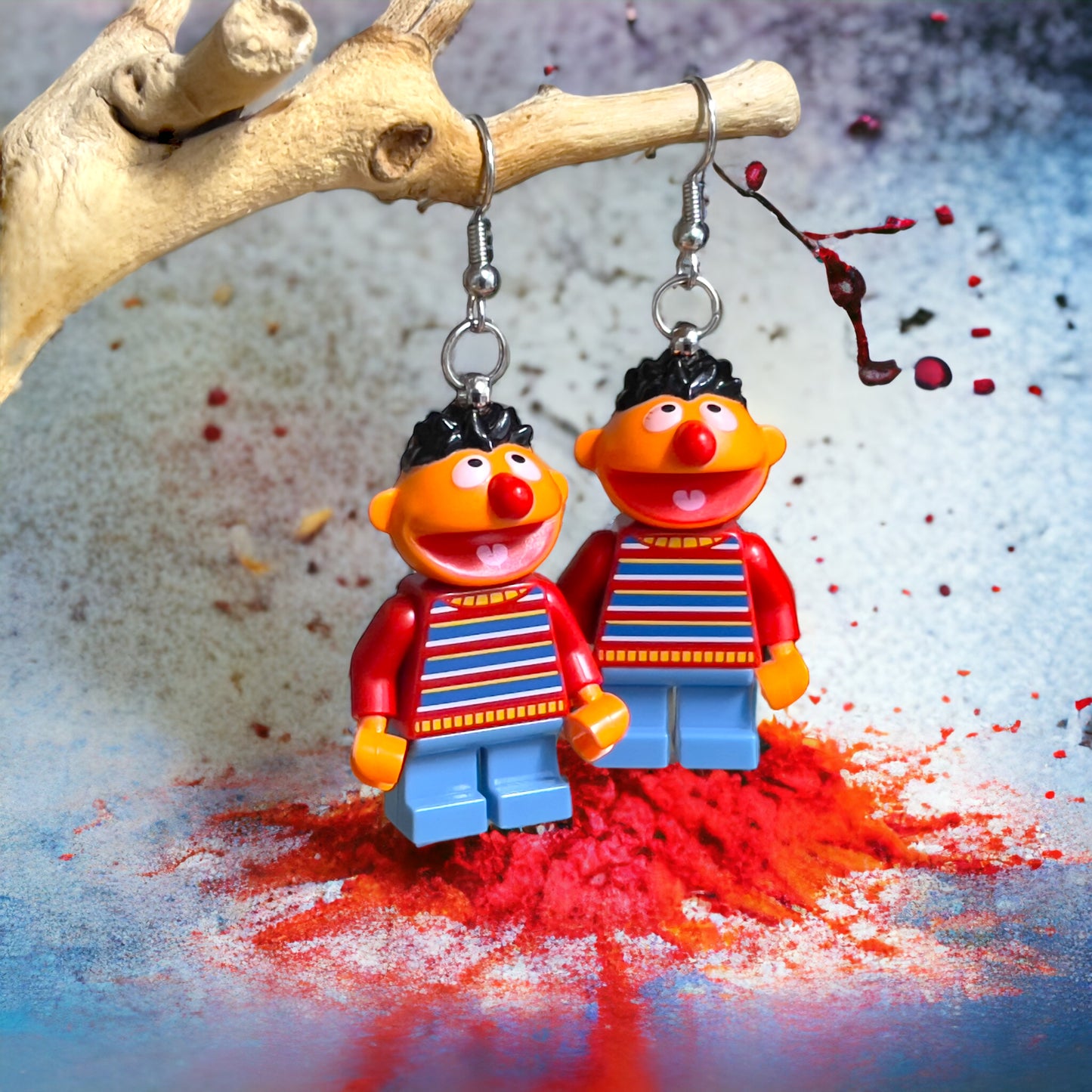 A lego minifigure character of the sesame street character Ernie wearing blue pants with a red hsirt that has blue, white, yellow stripes made into hook silver earrings hanging off a branch with a white backgroun red paint drops and red powder at the bottom.