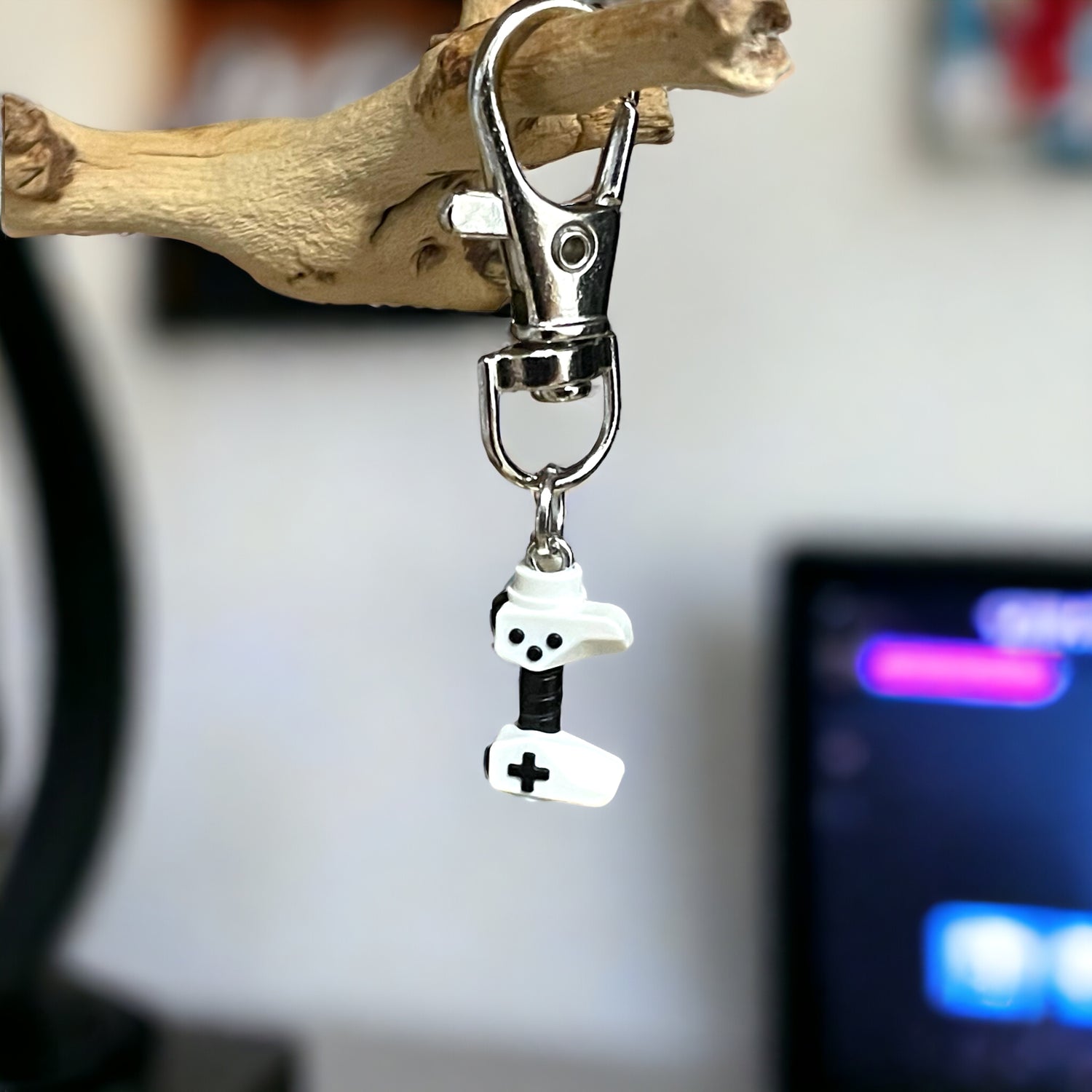 A white video game controller with black handle attached to a keychain clip hanging off a branch with a tv in the background