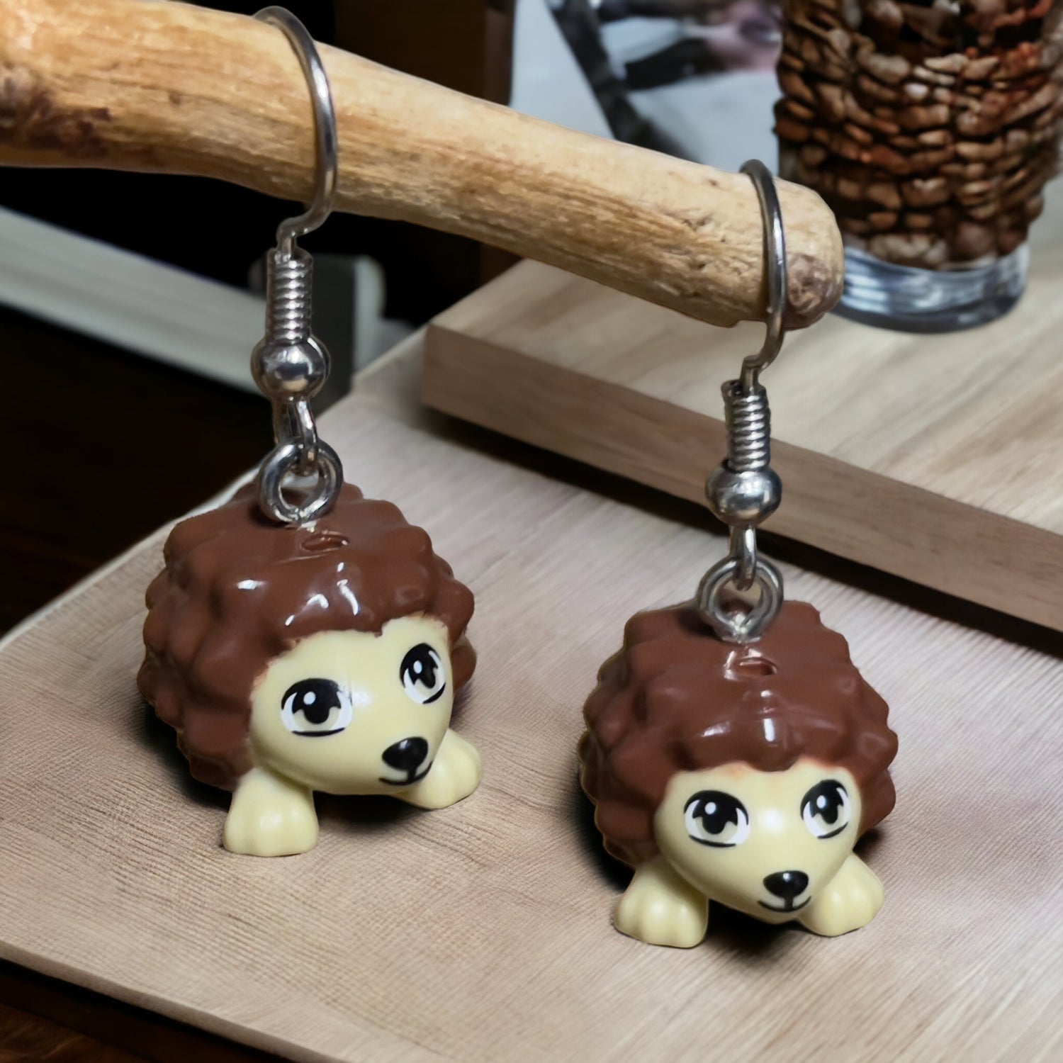 Two little brown lego hedgehogs with cream faces and black eyes and nose made into silver earrings hanging off a branch on a wooden table top with a vase in the background