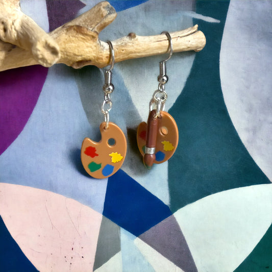 Two lego hook earrings set with a brown paint palettes with splashes of yellow, green, blue and red paint on it, one with a brown paintbrush with green tip hanging off a wooden branch with a geometric painting as a background