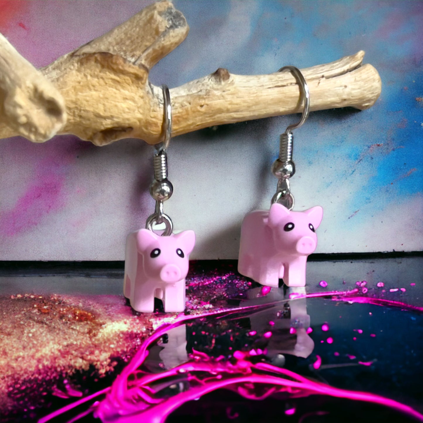 Two tiny pink piglets with hook earrings hanging off a branch with a colour pink and blue splash background and bright pink paint splash across the front of the picture with pink glitter on the floor