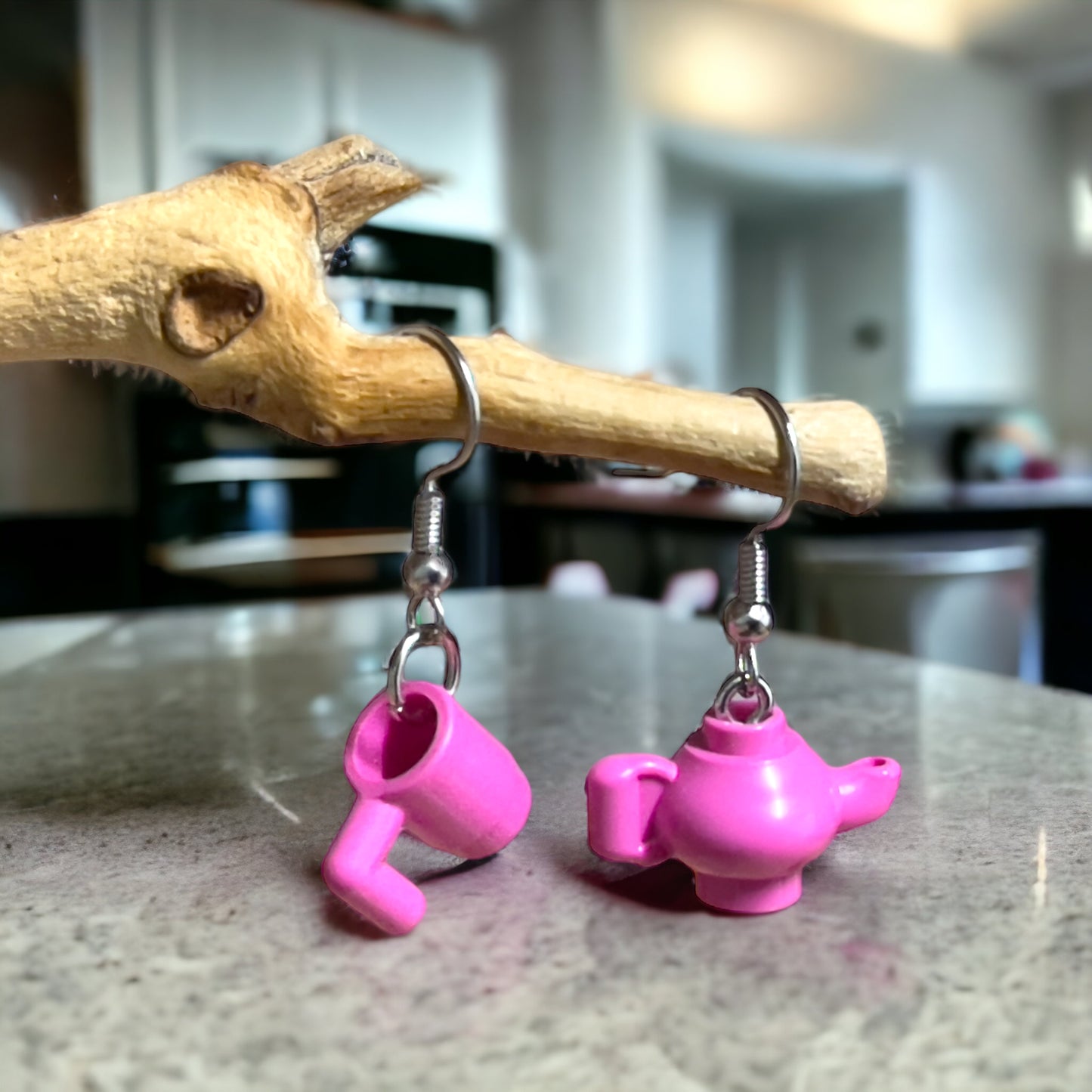 Pink lego tea pot and pink lego coffee cup with handle made into silver loop earrings hanging off a branch on a marble kitchen bench with a kitchen scene in the background