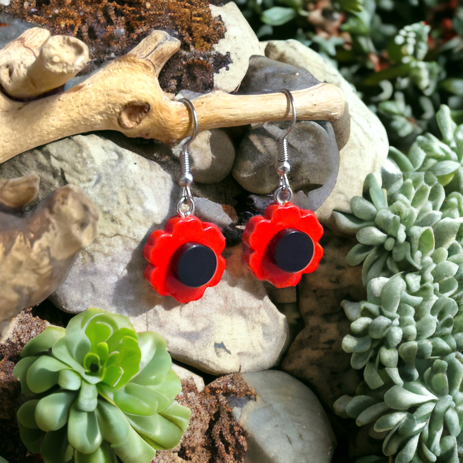 two red lego flowers with a black centre made with silver hook earrings hanging off a branch with rocks and green succulents in the background