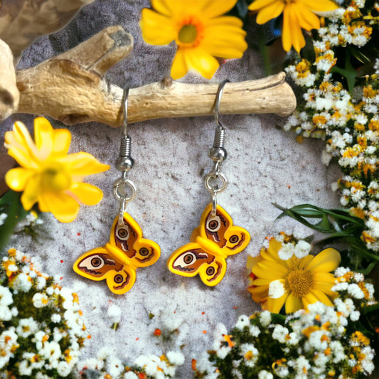 Two yellow lego butterflies with brown & white printing on wings on a set of silver hook earrings hanging off a branch with a background of yellow and white flowers with green stems and leaves.