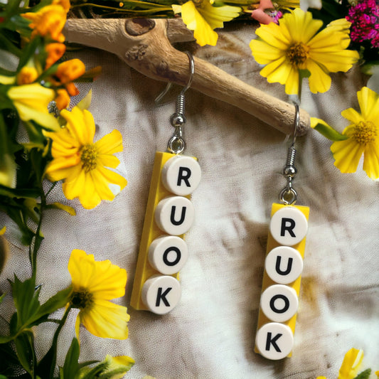 A canvas background with yellow flowers on the outside border of the picture, two hook style earrings made of a lego plate with the letters R U O K in white and black stuck to the lego plate