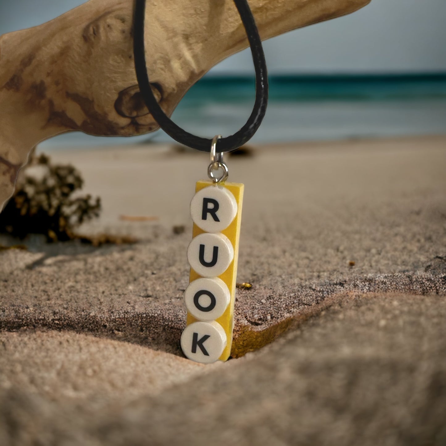 A black necklace with a yellow lego brick with lego round white dots with the letters R U OK in black hanging off a branch on sea sand with the blue ocean in the background