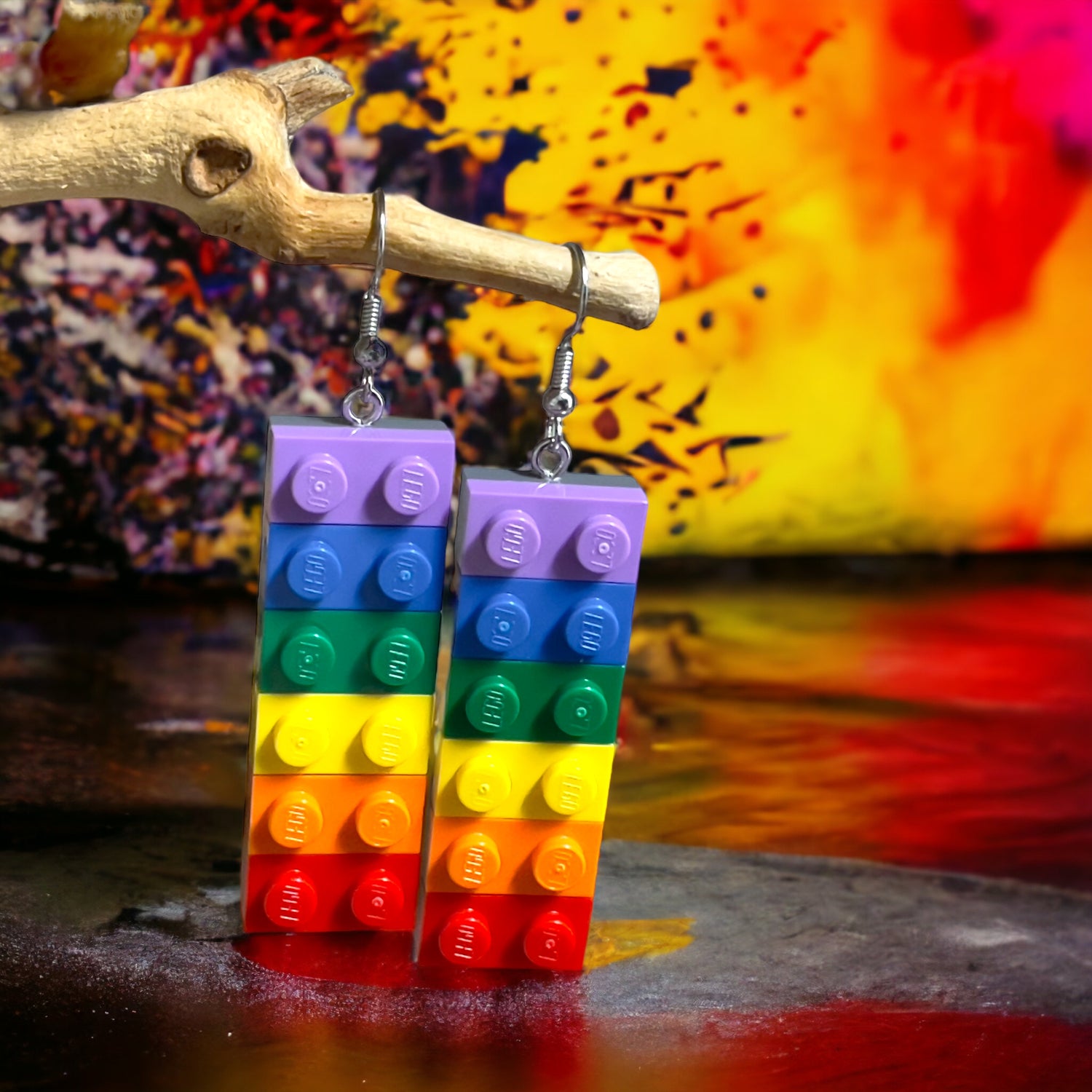 A pair of earrings made using lego 1x2 bricks in the colours of thre rainbow with purple at the top handing off a branch with a explosion of colour in the background