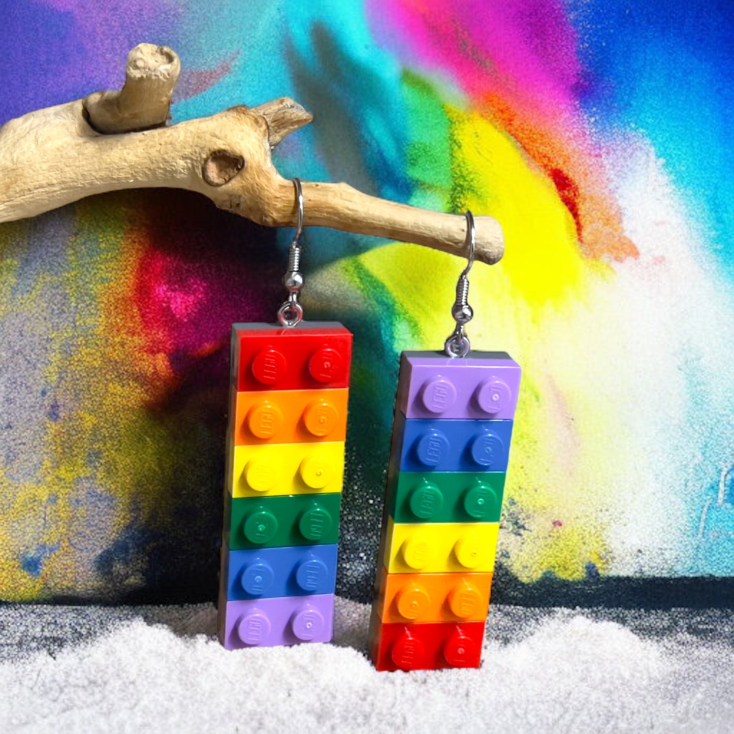 A pair of earrings made using lego 1x2 bricks in the colours of thre rainbow with one starting with purple at the top and the other with red at the top handing off a branch with a explosion of colour in the background