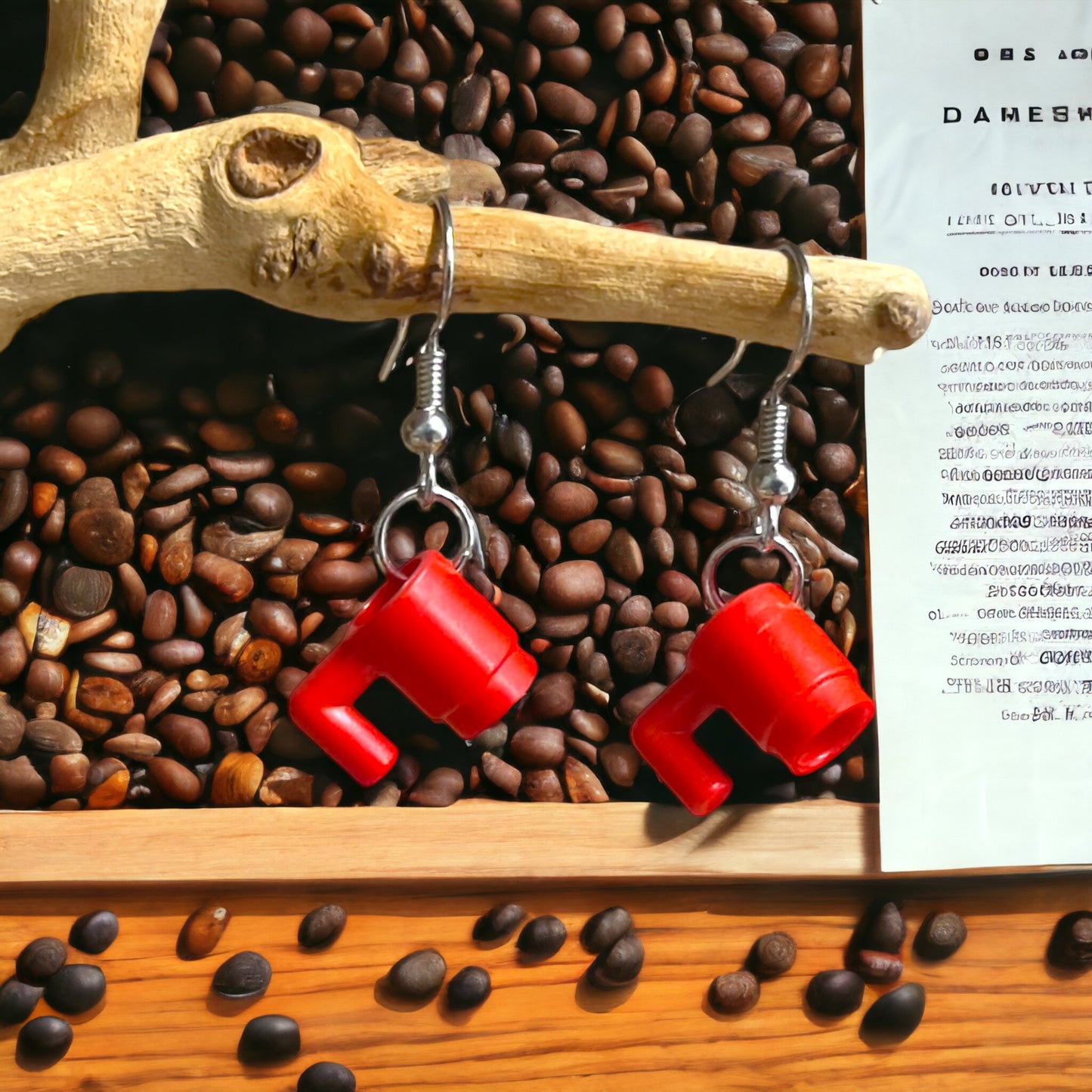 Two red lego coffee cups made into silver hook earrings hanging off a branch with coffee beans in the background and a page of a book on the right hand side