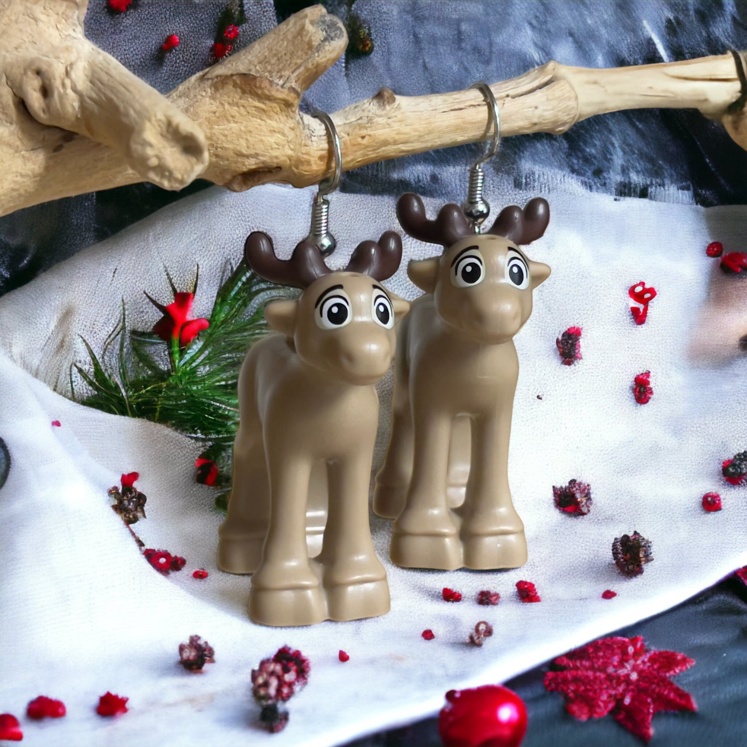 Two cute comic looking tan colour baby reindeer with brown antlers with silver hook earrings hanging over a branch with a white cloth sprinkled with red decorations and a little mistletoe in the background.