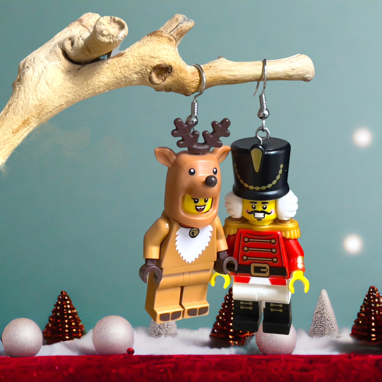 A light brown reindeer costimer with lego man head and a nutcracker costimer with lego man head made into silver loop earrings hanging off a branch with a light blue background and red velvet ground white baubles and red and gold pine cones with silver xmas trees