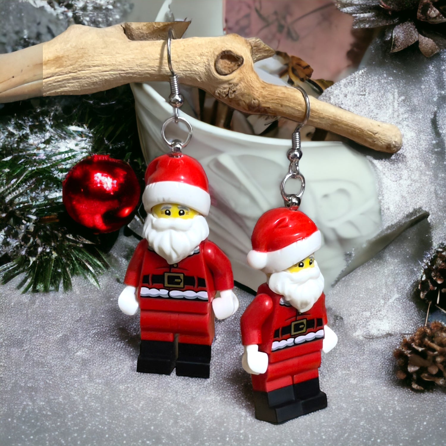 A lego minifigure of two santa's with a red suit and white beard, white gloves and black boots as hook earrings hanging off a branch in some green pine needles, a red round decoration and whites snow