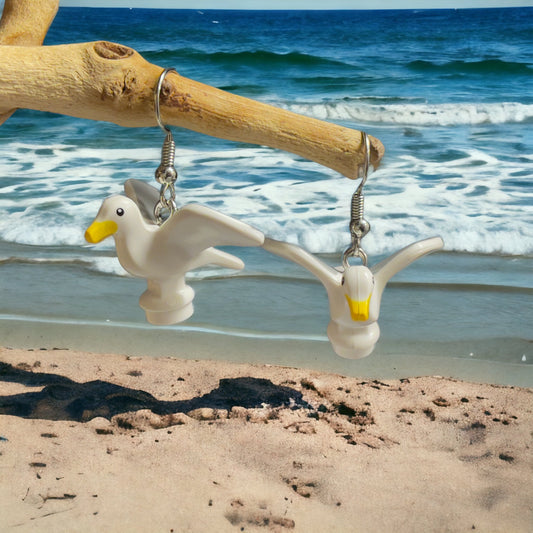 Two white lego seagull birds with yellow beaks makde into a silver hook earring hanging off a wooden branch with the white capped waves over blue ocean in the background and light brown sand at the bottom of the photo