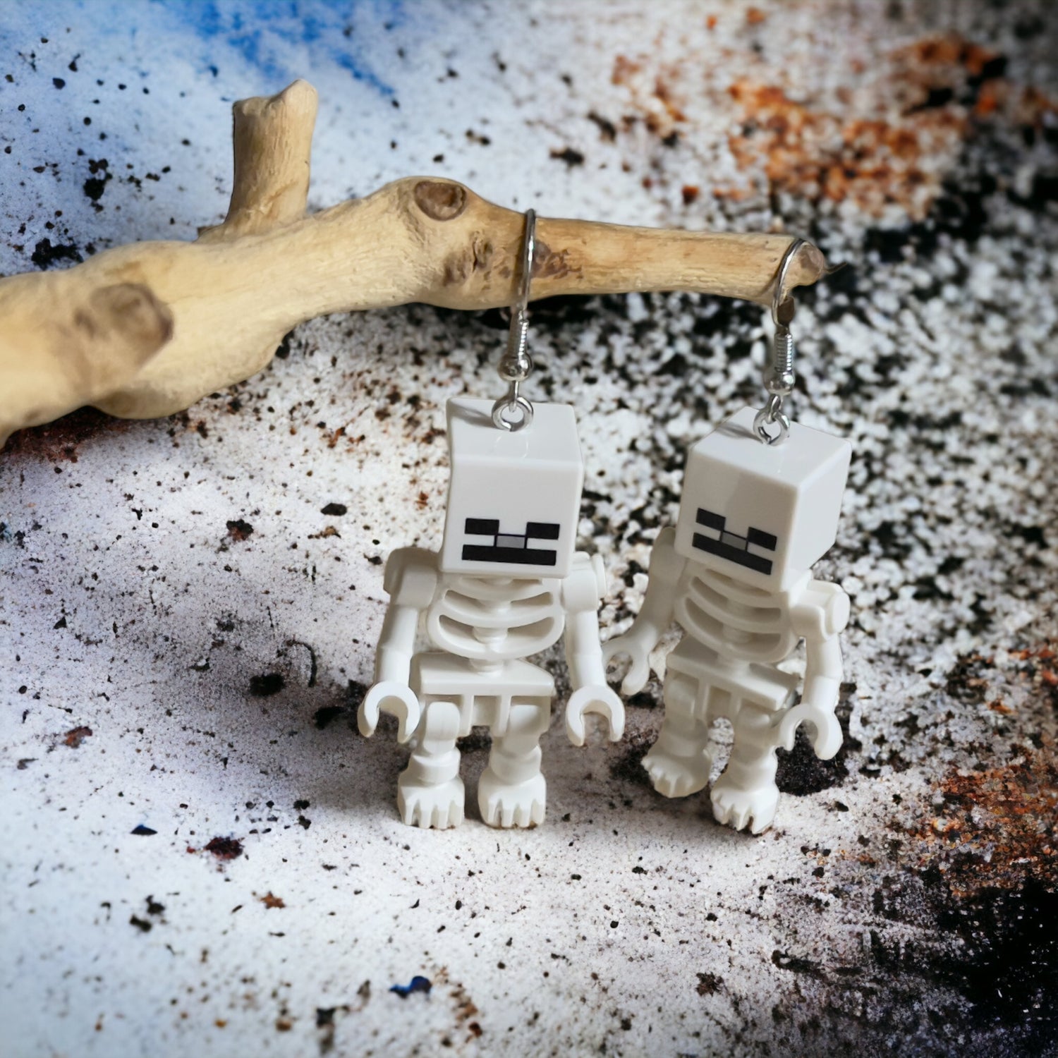 Two white skeletons with square heads and black face with silver hook earrings hanging on a branch with a black paint splattered white background 