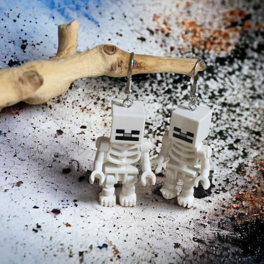 Two white skeletons with square heads and black face with silver hook earrings hanging on a branch with a black paint splattered white background 