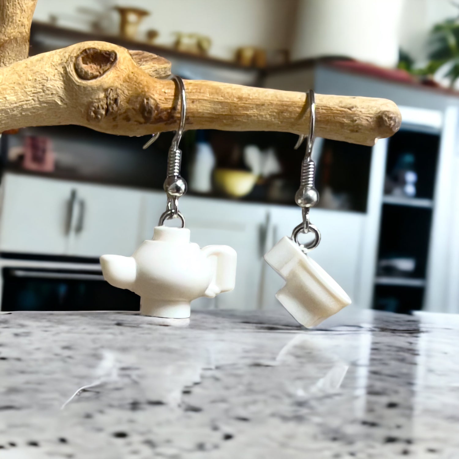 a white lego tea pot and white lego tea cup with handle made into lego silver loop earrings handing off a branch on a marble kitchen bench with kitchen shelves and cupboards in the background