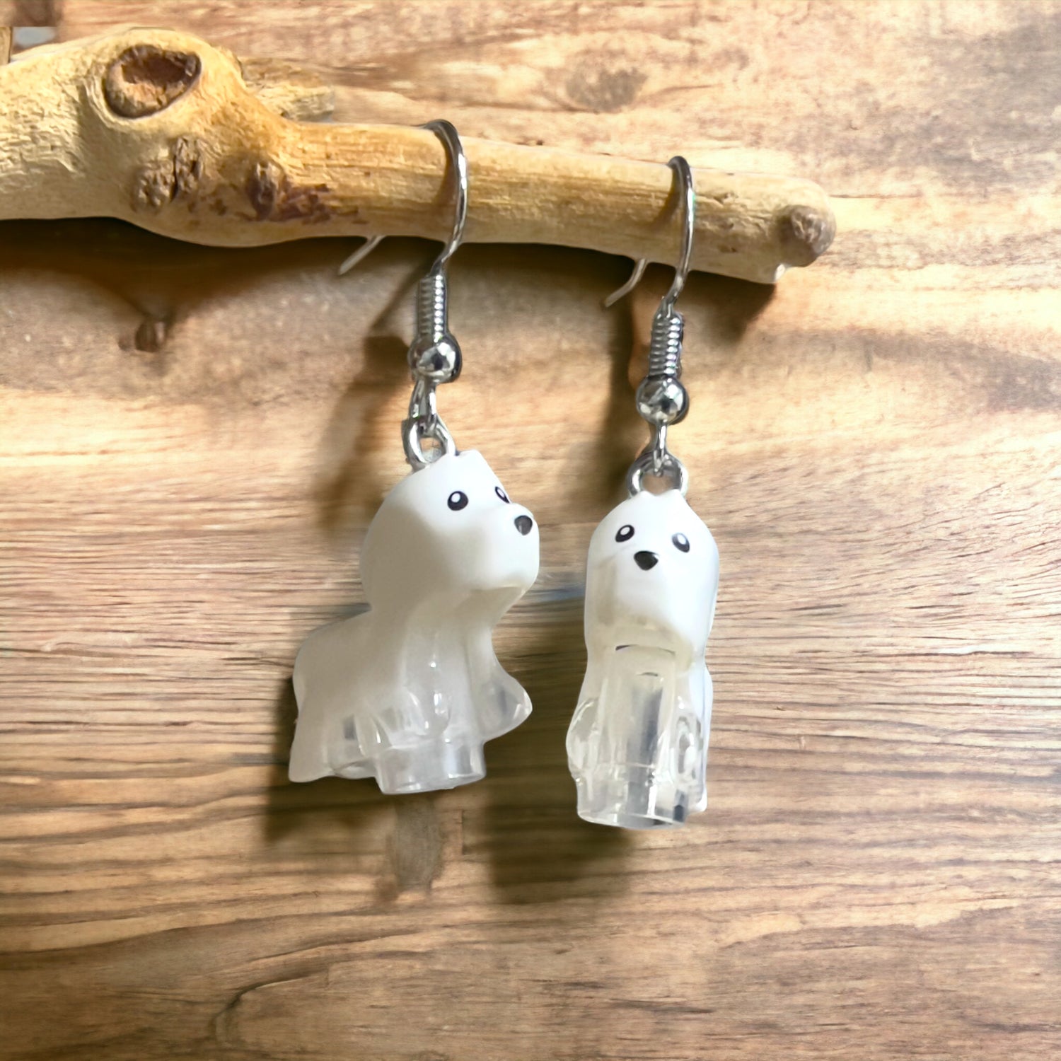 Two white little dogs with black eyes and nose made into silver hook earrings hanging off a branch with a light brown wooden background