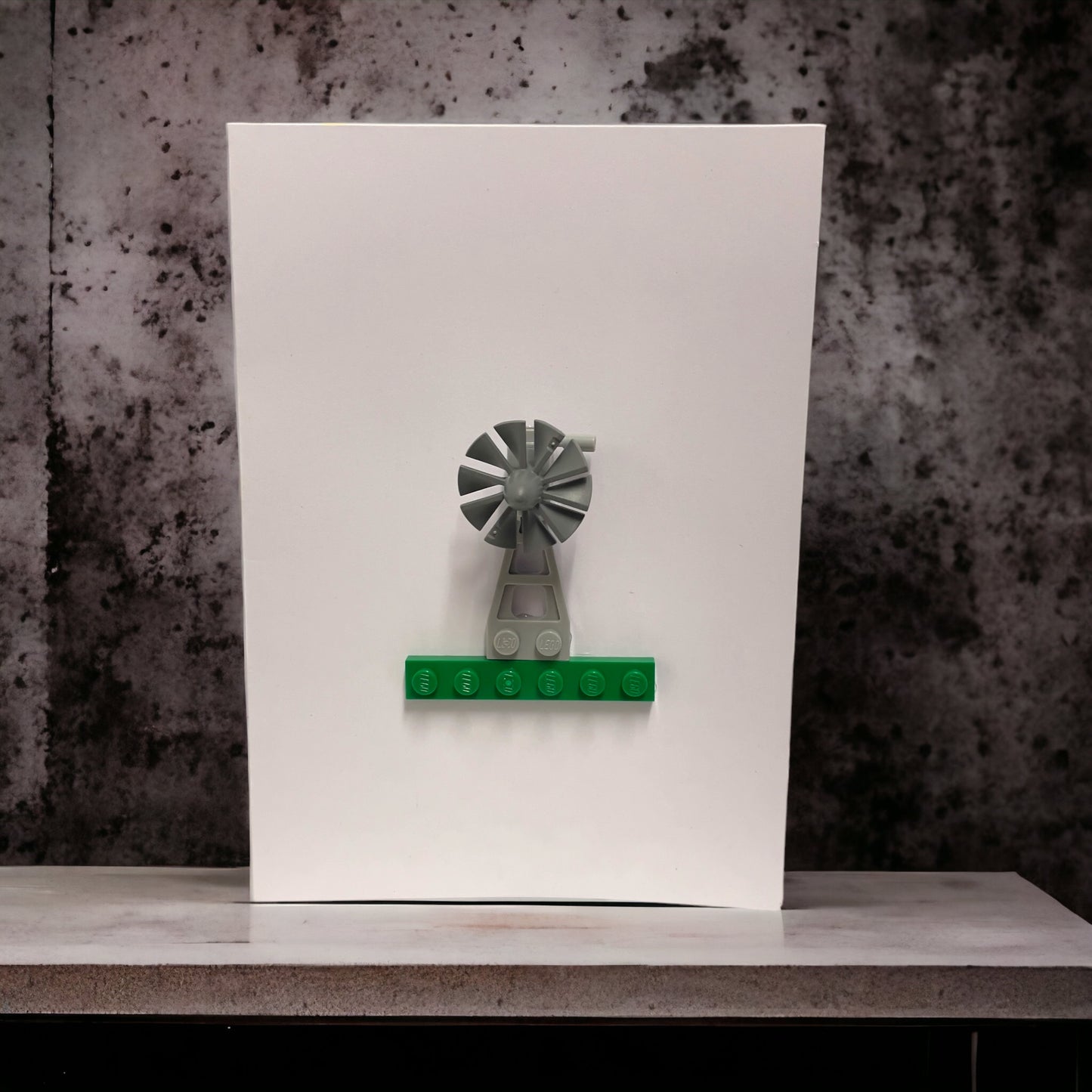 A grey lego windmill made from lego pieces on a green lego brick on a white blank greeting card sitting on a marble shelf with a marble background.