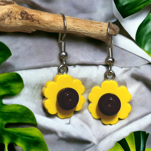 A pair of simple yellow flowers with a brown circle center as hook earrings. They are hooked on a branch that is resting on a light grey fabric background with green leaves laying flat in three corners of the image.