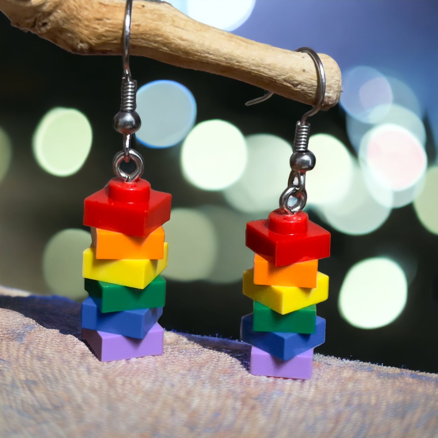 A pair of earrings made using lego 1x1 square studs in the colours of the rainbow with red at the top handing off a branch with a twinkling lights in the background