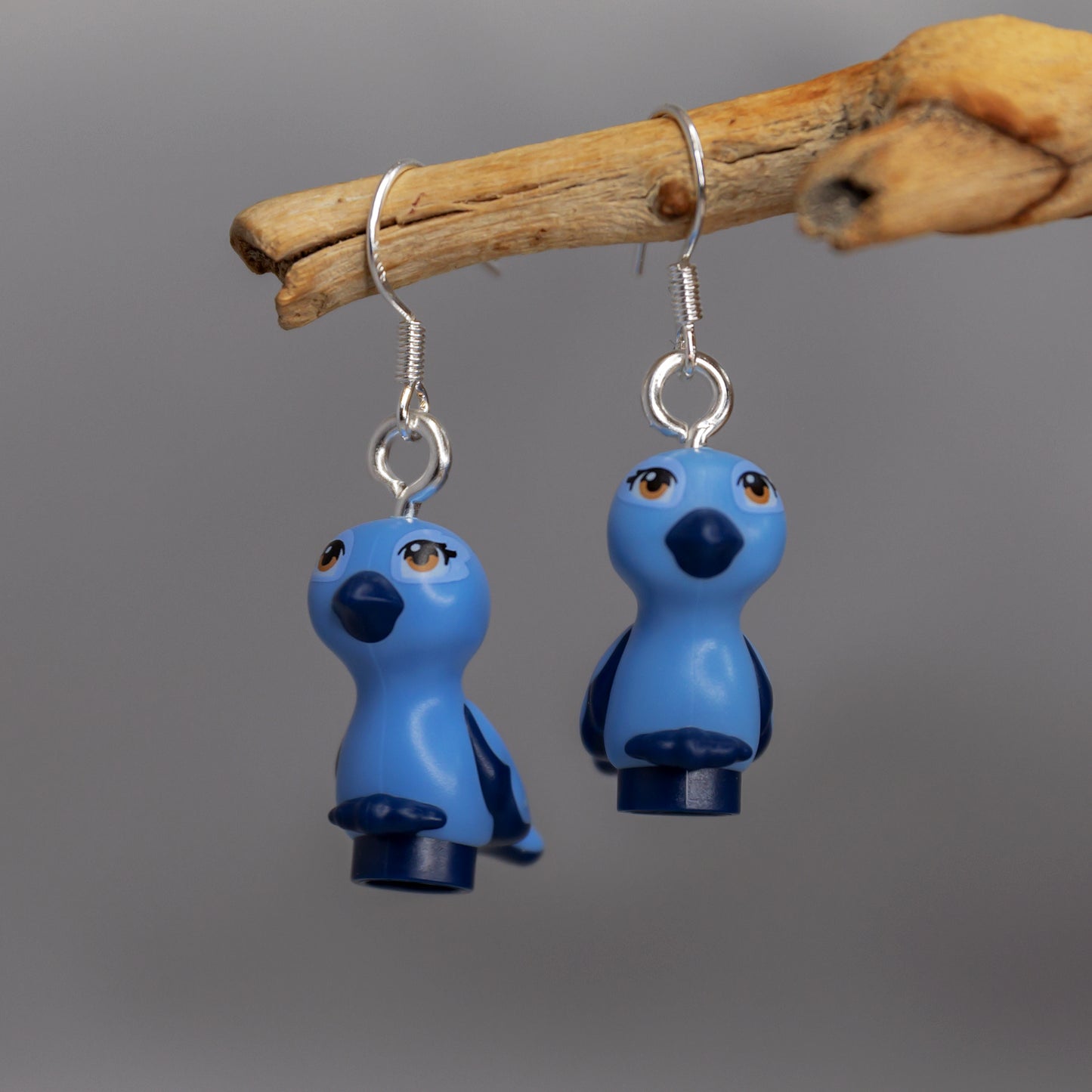 A pair of blue light blue birds with dak blue beaks , feet and wings with brown eyes made into silver earrings hanging off a branch with a grey background