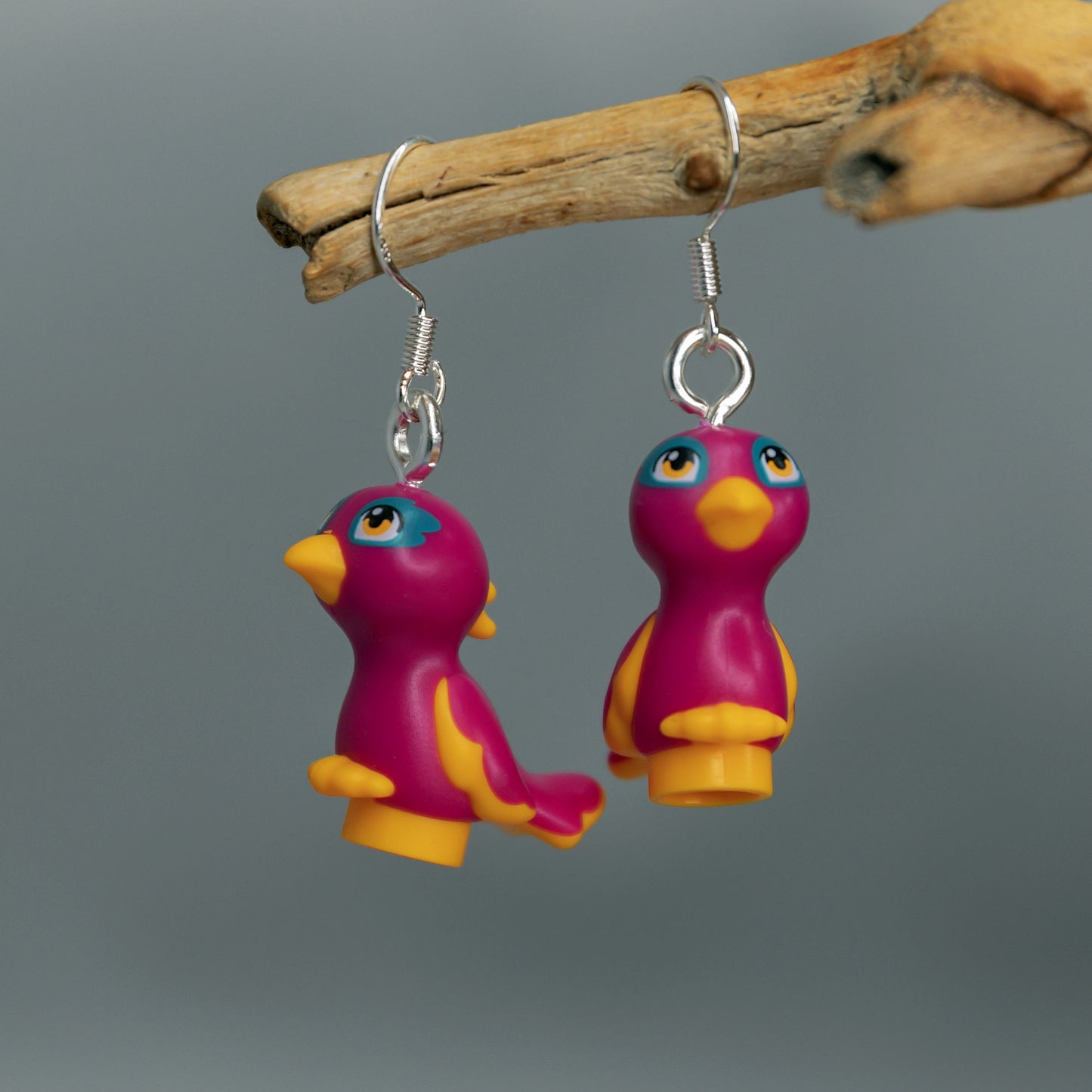 A pair of pink birds with yellow beaks , feet and wings with yellow eyes made into silver earrings hanging off a branch with a grey background